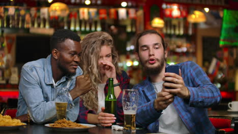 Two-men-and-a-woman-look-at-the-phone-screen-while-sitting-in-a-bar-and-prepare-to-take-a-shared-selfie.-Cheerful-youth-company-at-the-bar.